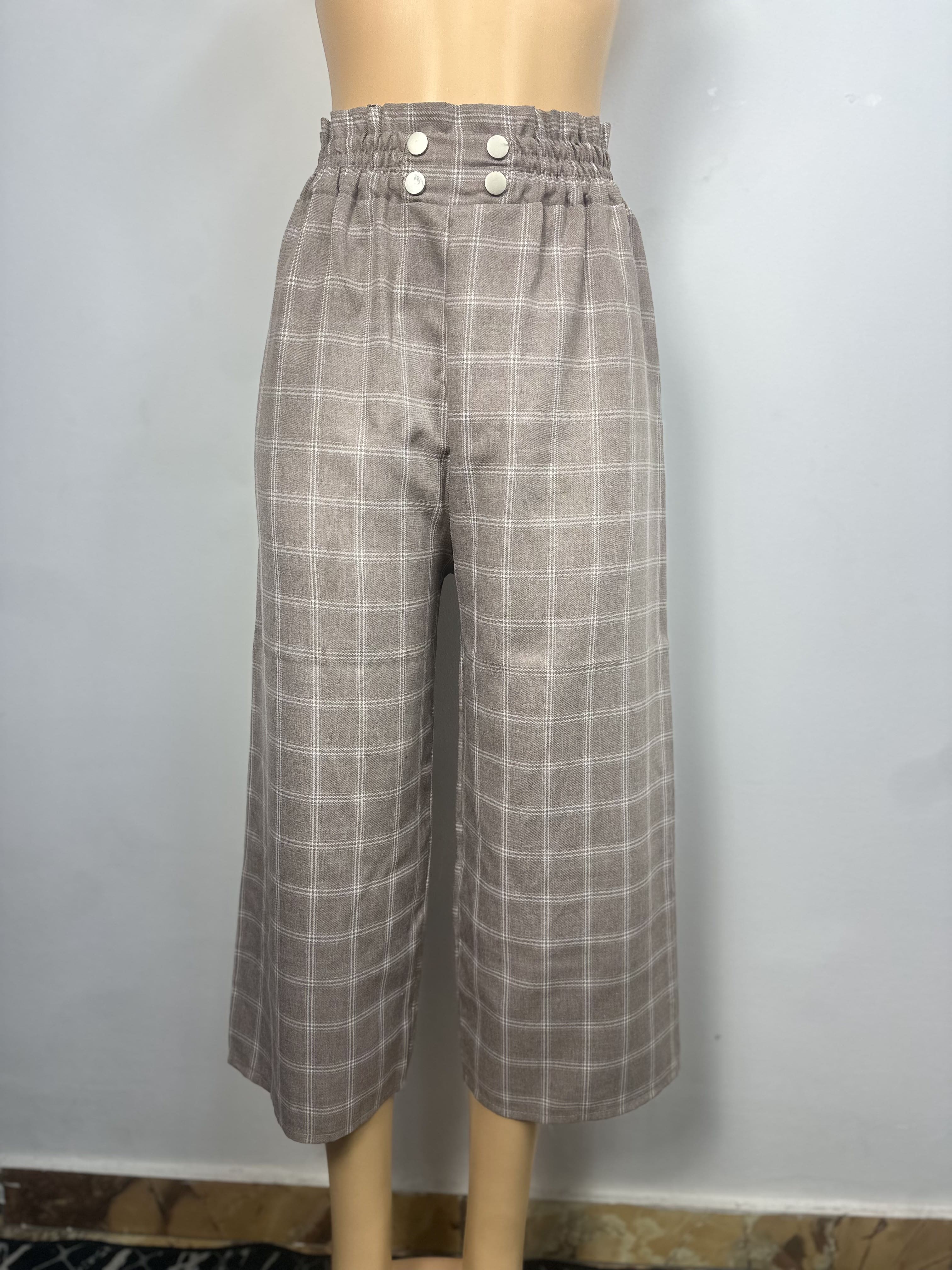 Pantalon  Gris-brun, à carreaux blancs, il est doté d'une ceinture élastique et de trois boutons clairs sur le côté gauche. Coupe ample et droite. L'arrière-plan est un mur gris clair uni avec une petite partie du sol à motifs visible.