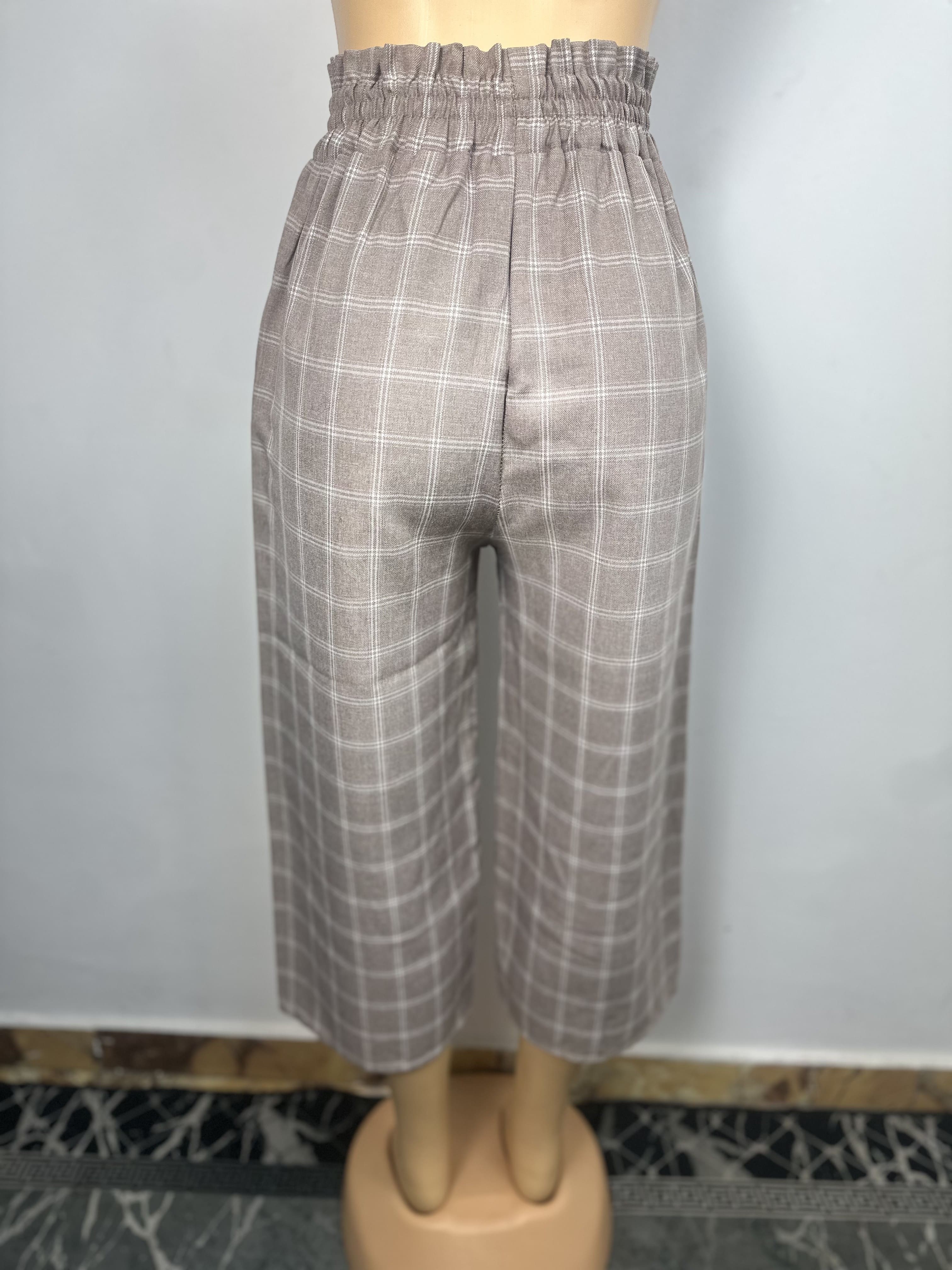 Pantalon  Gris-brun, à carreaux blancs, il est doté d'une ceinture élastique et de trois boutons clairs sur le côté gauche. Coupe ample et droite. L'arrière-plan est un mur gris clair uni avec une petite partie du sol à motifs visible.