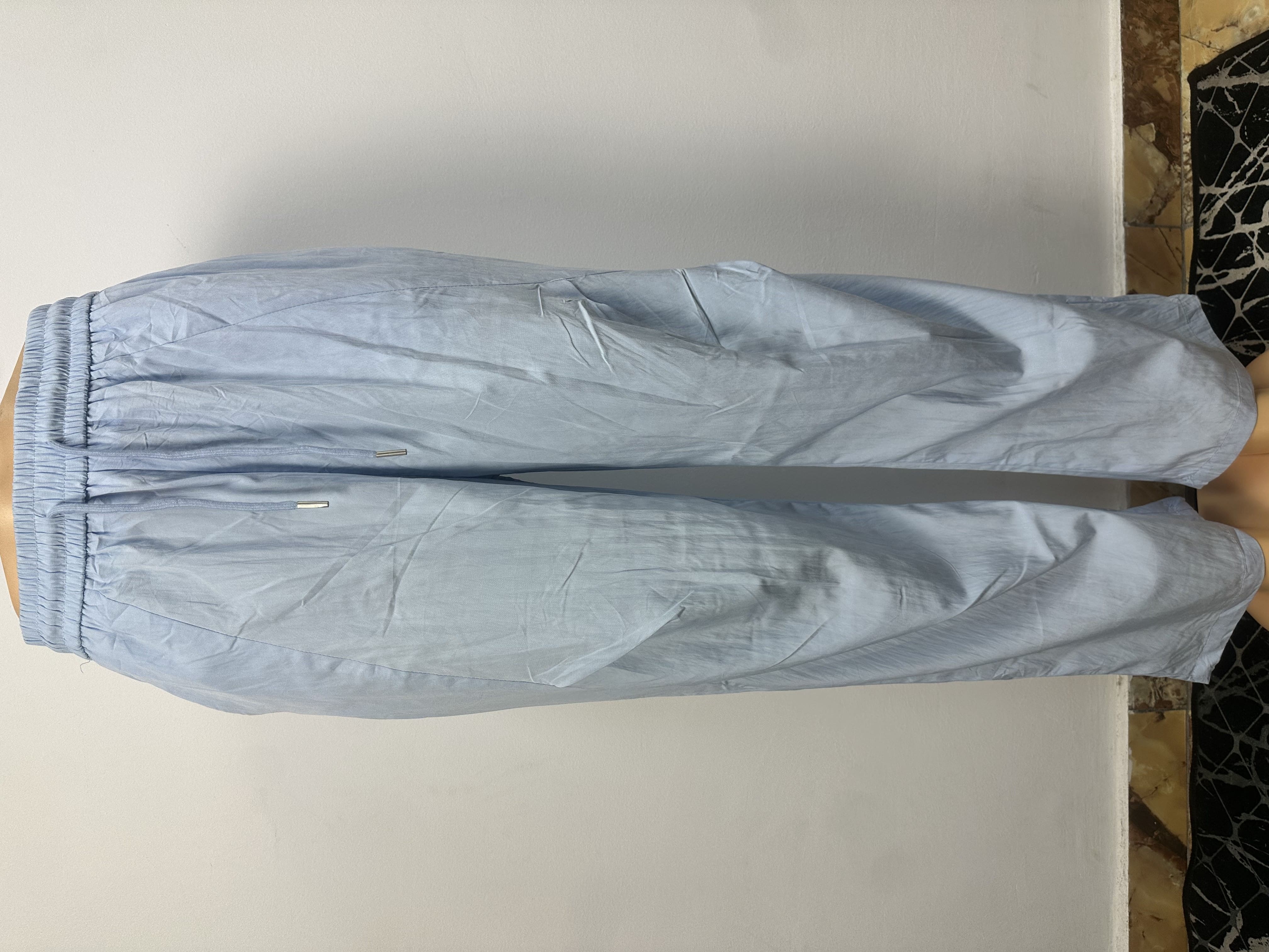 Pantalon bleu clair uni avec une taille élastique sans boutons ni cordon de serrage visibles. Coupe ample et légèrement fuselée. L'arrière-plan est un mur gris clair uni avec une petite partie du sol à motifs visible.