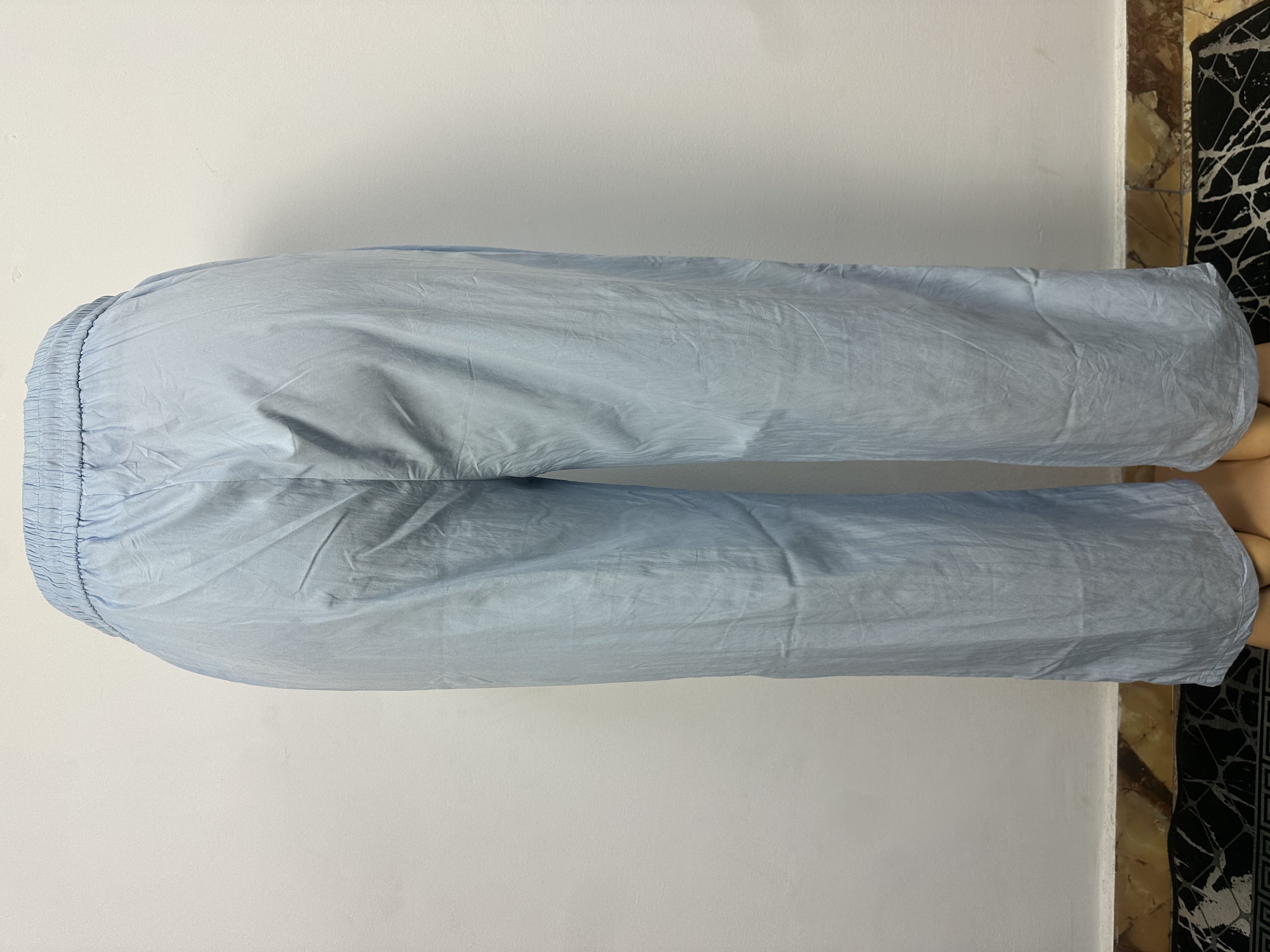 Pantalon bleu clair uni avec une taille élastique sans boutons ni cordon de serrage visibles. Coupe ample et légèrement fuselée. L'arrière-plan est un mur gris clair uni avec une petite partie du sol à motifs visible.