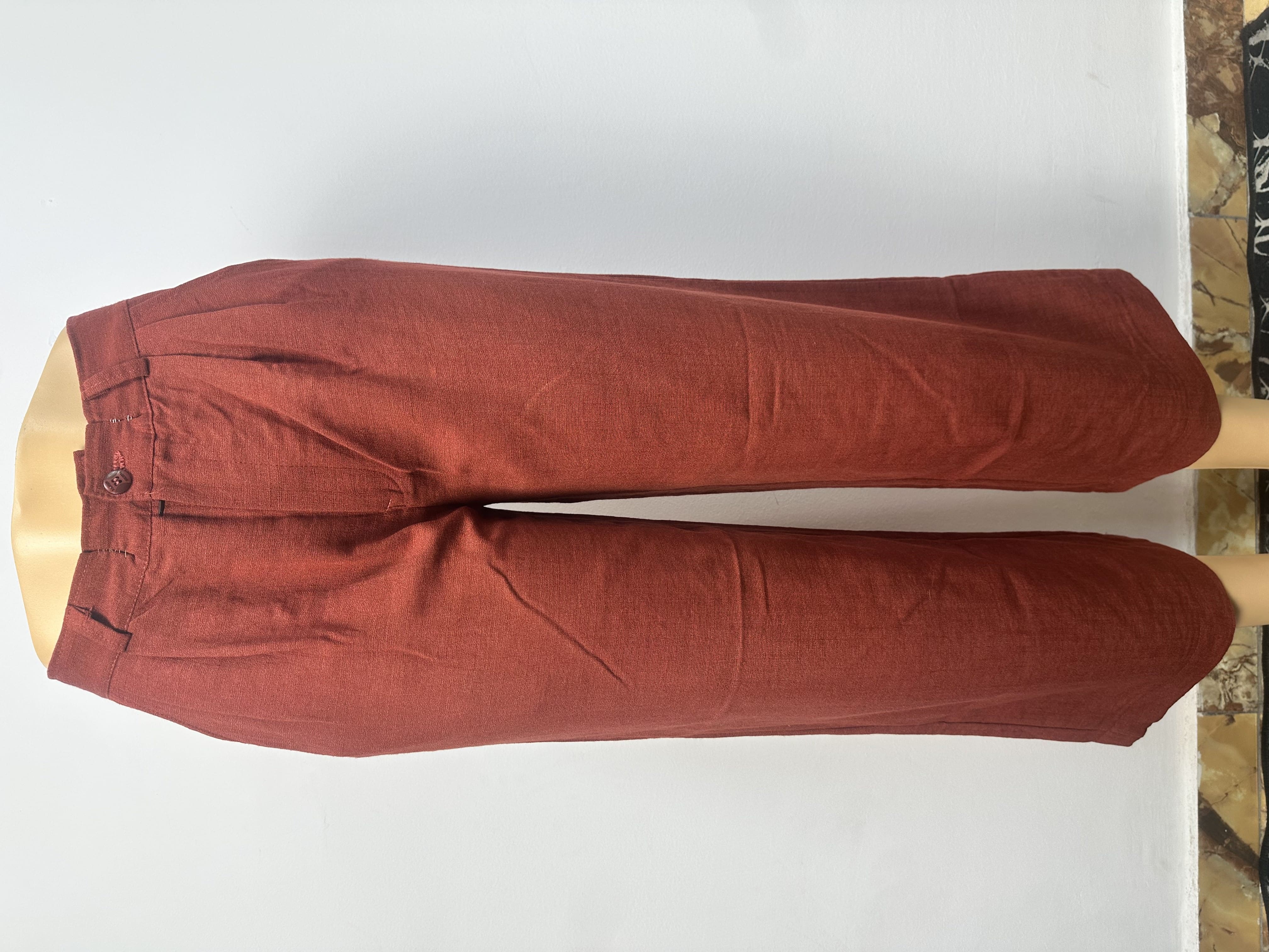 Pantalon rouge brique uni, avec une ceinture ajustée à passants et une fermeture boutonnée. La coupe est droite et ample, coupée au-dessus de la cheville.