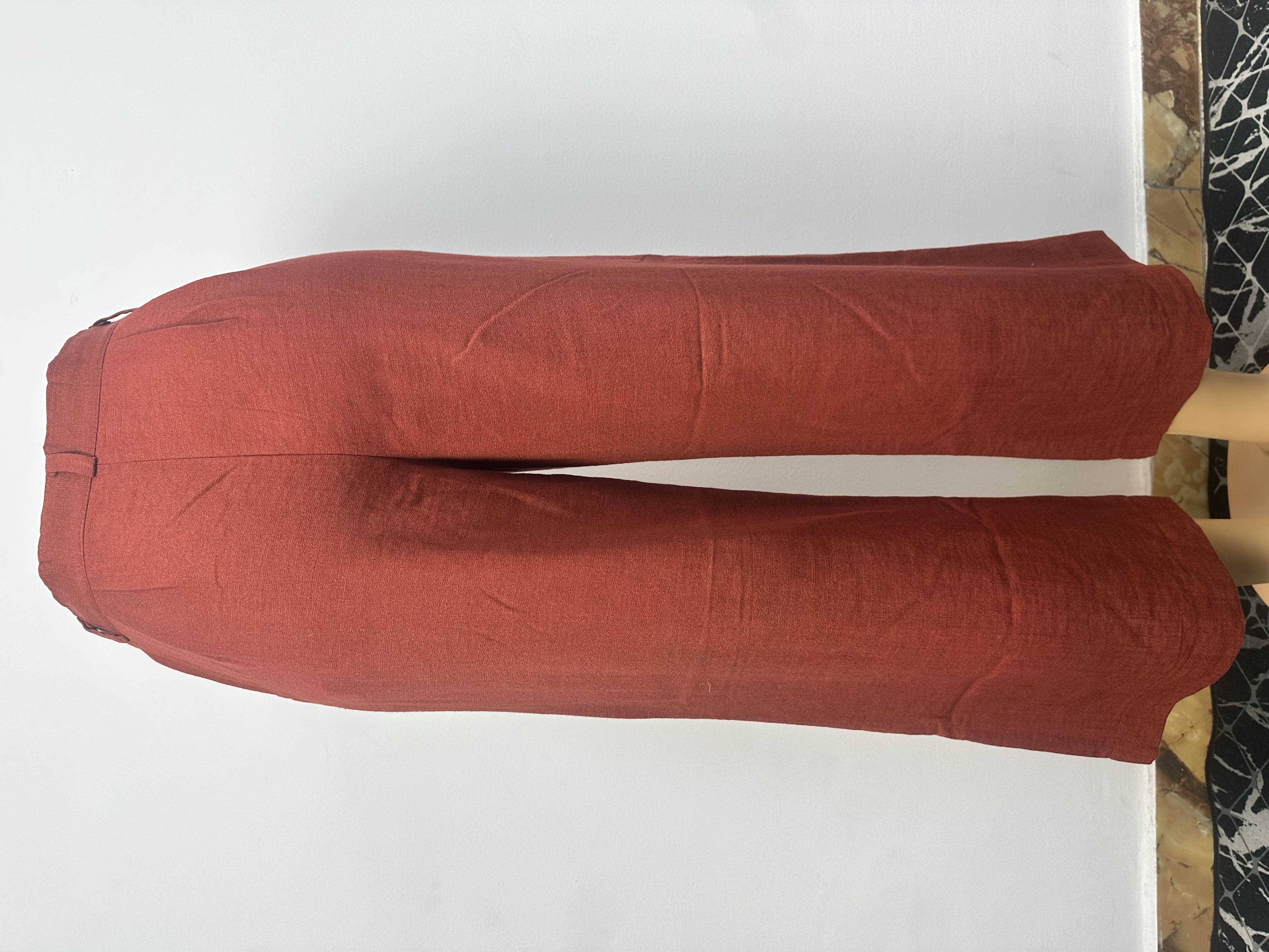 Pantalon rouge brique uni, avec une ceinture ajustée à passants et une fermeture boutonnée. La coupe est droite et ample, coupée au-dessus de la cheville.